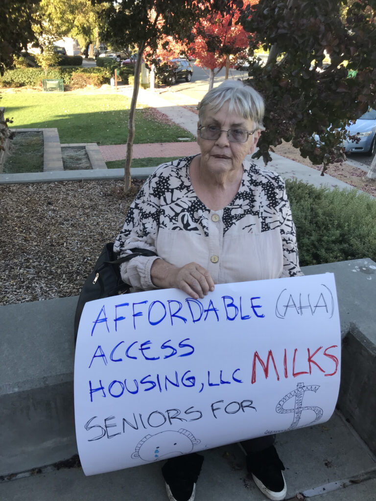 A senior woman holding a protest sign saying "Affordable access housing, LLC milks seniors for $"