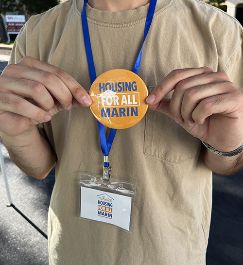 Photo of a man holding a Housing For All Marin logo button. The button is orange with blue and white lettering.